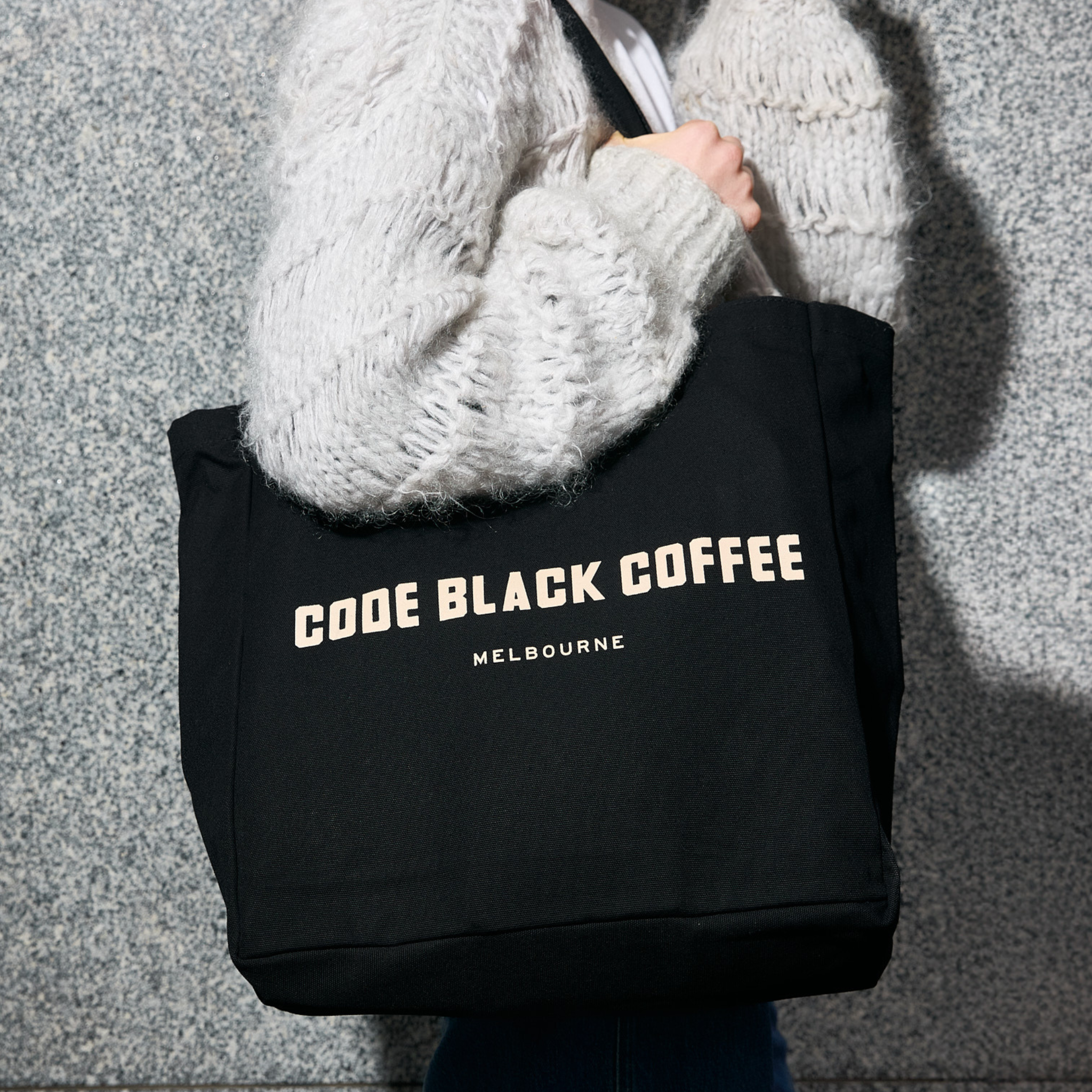 Person holding a black tote bag with 'Code Black Coffee Melbourne' text against a gray background