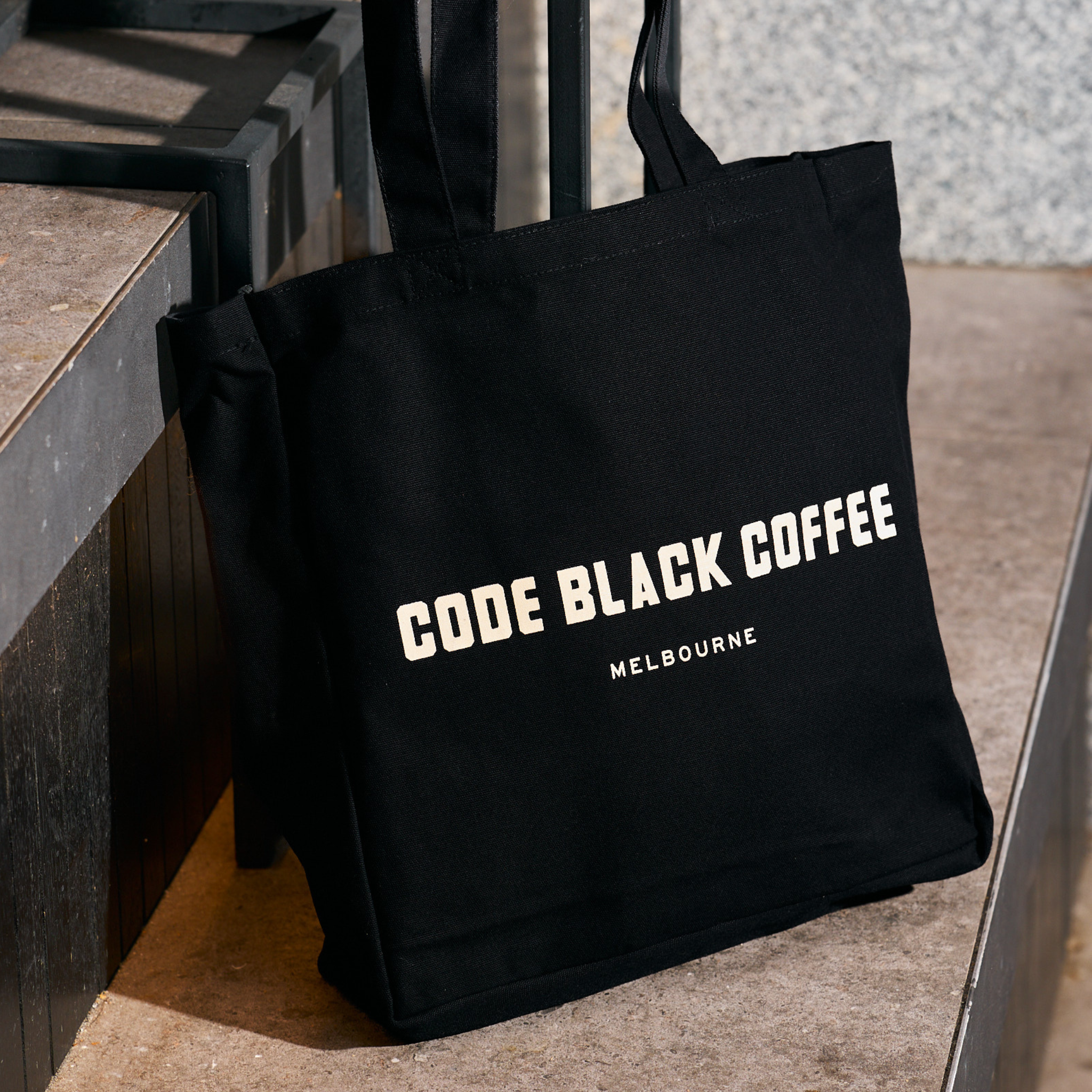 Black tote bag with 'Code Black Coffee Melbourne' text on a stone surface.
